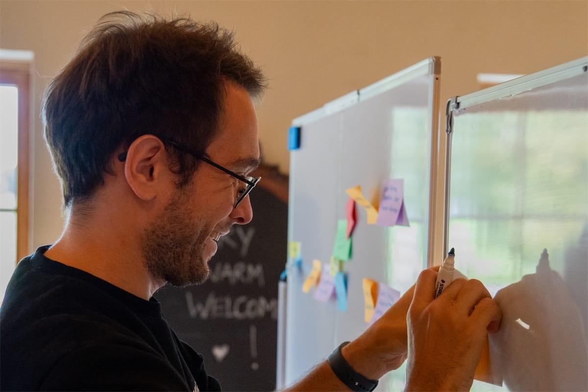 Ein Kollege klebt beim Event Storming lachend eine Haftnotiz an ein Whiteboard